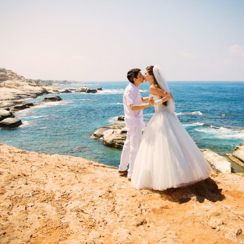 Elegant smiling bride and groom walking on the beach, kissing and having fun, wedding ceremony near the rocks. Mediterranean Sea, Cyprus